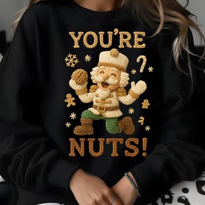 May include: Black sweatshirt with a knitted design of a nutcracker soldier. The text "YOU'RE NUTS!" is in a gold color. The design includes snowflakes and gingerbread men. A fun, festive holiday-themed top.