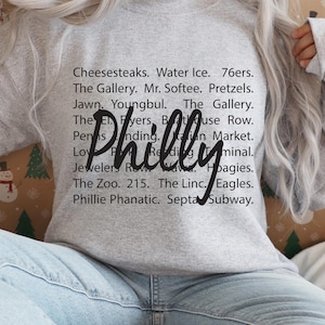 May include: A gray sweatshirt with the word "Philly" written in large, black cursive letters. The sweatshirt has a list of Philadelphia-related words and phrases printed on it, including "Cheesesteaks", "Water Ice", "76ers", "The Gallery", "Mr. Softee", "Pretzels", "Jawn", "Youngbul", "The Gallery", "The El", "Flyers", "BatHouse Row", "Penn's Landing", "Italian Market", "Love Park", "Reading Terminal", "Jewelers Row", "The Zoo", "215", "The Linc", "Eagles", "Phillie Phanatic", and "Septa Subway".