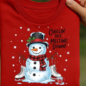Puede incluir: Camiseta roja de manga larga con un diseño de muñeco de nieve. El muñeco de nieve lleva un sombrero de copa negro, una bufanda a cuadros rojos y negros y patines de hielo. El texto de la camiseta dice "Chillin' Out, Melting Down!". Copos de nieve y una taza de chocolate caliente completan el diseño.
