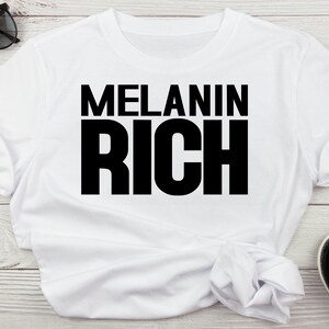 May include: White t-shirt with black text that reads "MELANIN RICH".