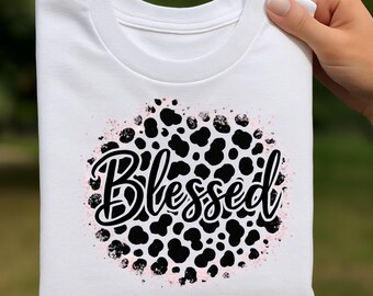 May include: White crew neck sweatshirt with a black and pink design. The design features black spots and the word "Blessed" in a cursive font. The design is surrounded by a pink speckled border.