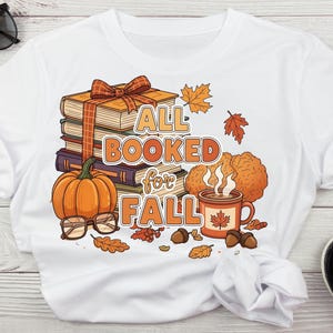 May include: White t-shirt with a colorful fall-themed graphic. The design includes a stack of books tied with a ribbon, a pumpkin, a coffee mug, and the text "ALL BOOKED for FALL". Autumn leaves and acorns complete the design.