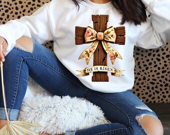May include: White sweatshirt with a brown wooden cross decorated with a floral bow and the text "He is Risen".