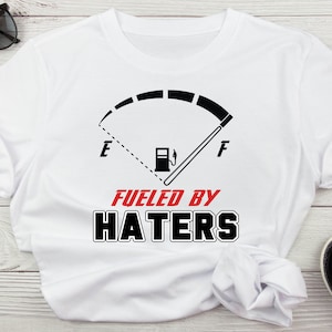 Puede incluir: Camiseta blanca con un gráfico de indicador de combustible en blanco y negro que dice "Fueled by Haters".