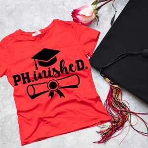 以下が含まれることがあります： 黒い文字で「PH.inished.」と書かれた赤いTシャツ。テキストには、黒い卒業帽と卒業証書が添えられています。