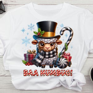 Op de afbeelding: Wit T-shirt met een cartoon schaap met een hoge hoed, monocle en sjaal, met een zuurstok. Het shirt heeft de tekst "BAA HUMBUG!" in rood en wit. Cadeaus en sneeuwvlokken zijn ook in het ontwerp verwerkt.