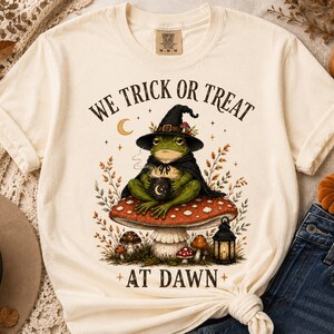 May include: A cream-colored t-shirt featuring a graphic of a green frog wearing a black witch hat and cape, sitting on a red and white mushroom. The frog holds a black mug with a crescent moon. Text reads WE TRICK OR TREAT AT DAWN.