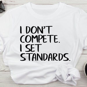 May include: White t-shirt with black text that reads "I don't compete. I set standards."