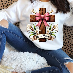 May include: White sweatshirt with a brown wooden cross with a pink bow and white calla lilies. The cross has the text "He is Risen" on it.