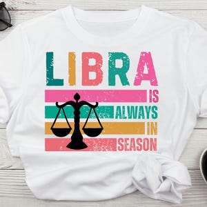 May include: White t-shirt with a colorful graphic design that says "LIBRA is ALWAYS in SEASON" with a stylized scale design.