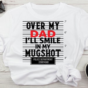 May include: White t-shirt with a black and red graphic that says "Over My Dad I'll Smile In My Mugshot" with a police department ID number below.