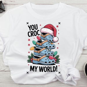 Puede incluir: Camiseta blanca con un gráfico de zuecos azules apilados con un gorro de Papá Noel, luces de Navidad y el texto "YOU CROC MY WORLD!". El diseño incluye estrellas rojas y acebo.