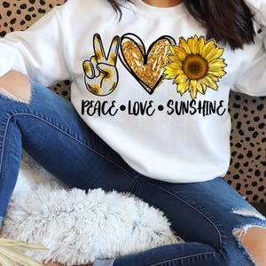 May include: White sweatshirt with a peace sign, heart, and sunflower graphic. The text "Peace Love Sunshine" is printed below the graphic.