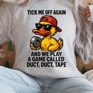 May include: A light gray sweatshirt featuring a cartoon duck wearing sunglasses and a red cap, holding duct tape. The text "TICK ME OFF AGAIN" and "AND WE PLAY A GAME CALLED DUCT, DUCT, TAPE" is printed above and below the duck illustration.