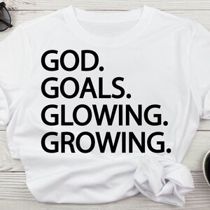 以下が含まれることがあります： 黒い文字で「GOD. GOALS. GLOWING. GROWING.」と書かれた白いTシャツ