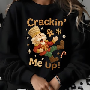 May include: Black sweatshirt with a festive design. The graphic features a cartoon nutcracker with the text "Crackin' Me Up!" in a playful font. The design includes gingerbread men, snowflakes, and a candy cane.
