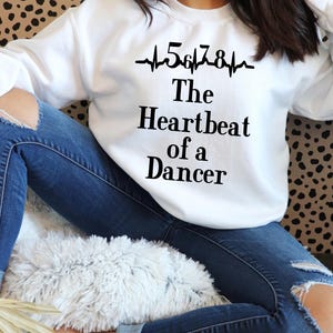 Könnte beinhalten: Weißer Pullover mit einem schwarzen Aufdruck, der "5678 The Heartbeat of a Dancer" mit einer stilisierten EKG-Linie lautet.