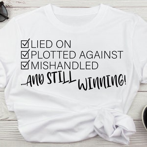 May include: White t-shirt with a black graphic that says "Lied on, Plotted Against, Mishandled...And Still Winning!" The graphic is a checklist with checkmarks next to each item.