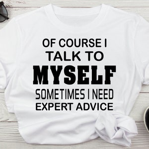 May include: White t-shirt with the text "OF COURSE I TALK TO MYSELF SOMETIMES I NEED EXPERT ADVICE" in black. The shirt is folded, with sunglasses and sandals in the background.