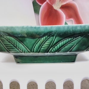 May include: A green ceramic planter with a square shape and a raised design of leaves around the rim.