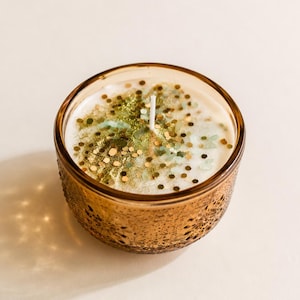May include: A scented candle in a round, amber-colored glass jar with an intricate, embossed floral pattern. The white wax surface is topped with a scattering of gold and green glitter particles and features a single, centered white cotton wick.