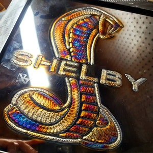 Puede incluir: Una obra de arte en metal que representa una cobra con la palabra "SHELBY". La cobra y el texto están en tonos dorados, naranjas, azules y morados, sobre un fondo metálico oscuro. La obra muestra detalles de soldadura intrincados.