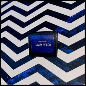 Könnte beinhalten: Eine schwarze und silberne Anstecknadel mit blauem Hintergrund und Glitzer. Der Text auf der Anstecknadel lautet "Directed by David Lynch".