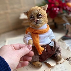 May include: A handmade plush toy cat with a white sweater, orange scarf, and brown corduroy pants. The cat has a distressed, vintage look and is sitting on a book.