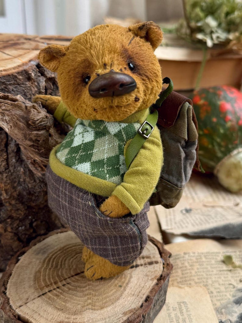 Puede incluir: Un oso de peluche marr&oacute;n que lleva un su&eacute;ter verde y blanco, pantalones marrones y una mochila marr&oacute;n. El oso est&aacute; de pie sobre un tronco de madera.