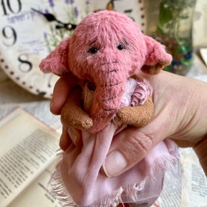 May include: A small pink elephant teddy bear wearing a pink dress with a ruffled trim. The bear has a long trunk and sad-looking eyes.