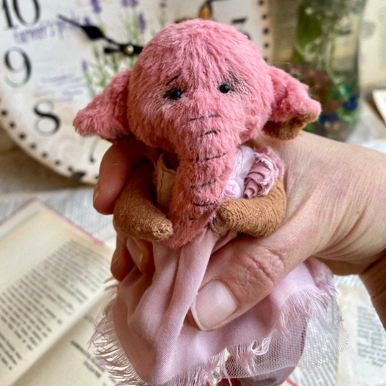 May include: A small pink elephant teddy bear wearing a pink dress with a ruffled trim. The bear has a long trunk and sad-looking eyes.