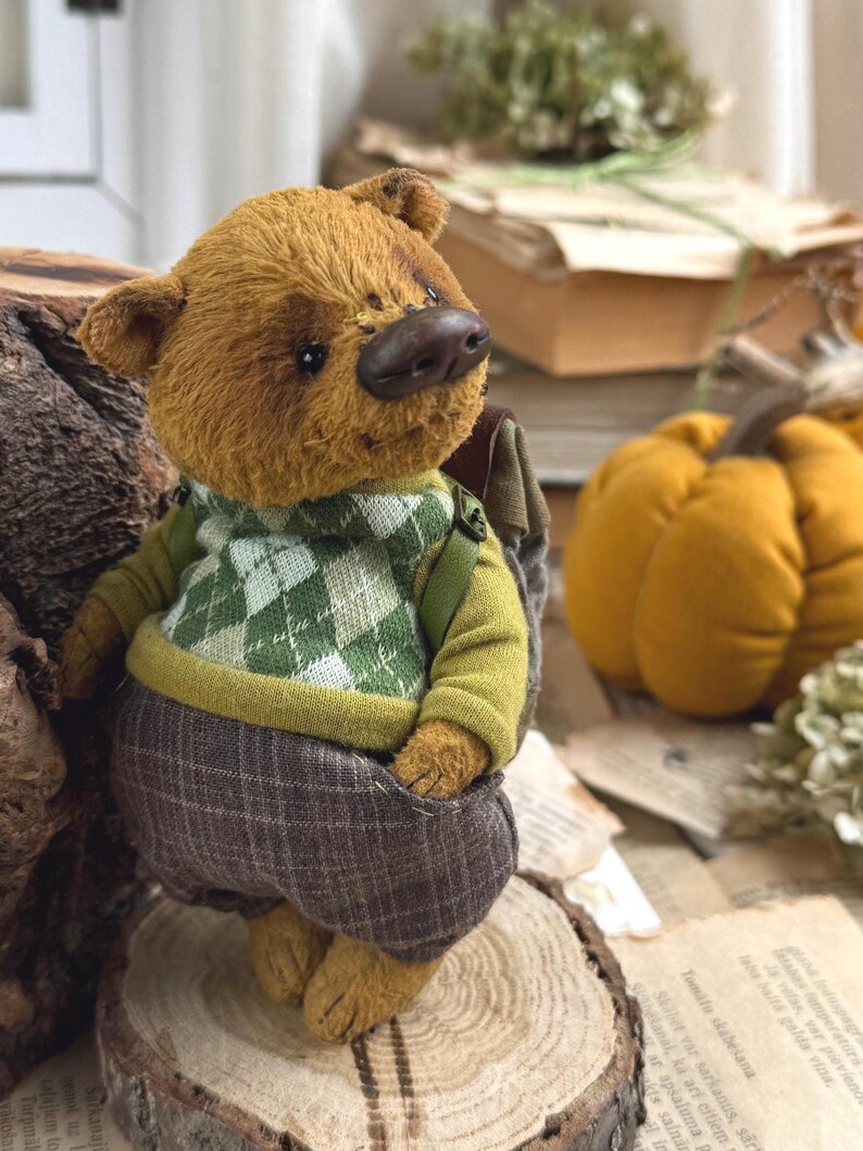 Puede incluir: Un peque&ntilde;o oso de peluche marr&oacute;n que lleva un su&eacute;ter verde y blanco y pantalones de cuadros marrones. El oso est&aacute; de pie sobre un tronco de madera y lleva una peque&ntilde;a mochila a la espalda.