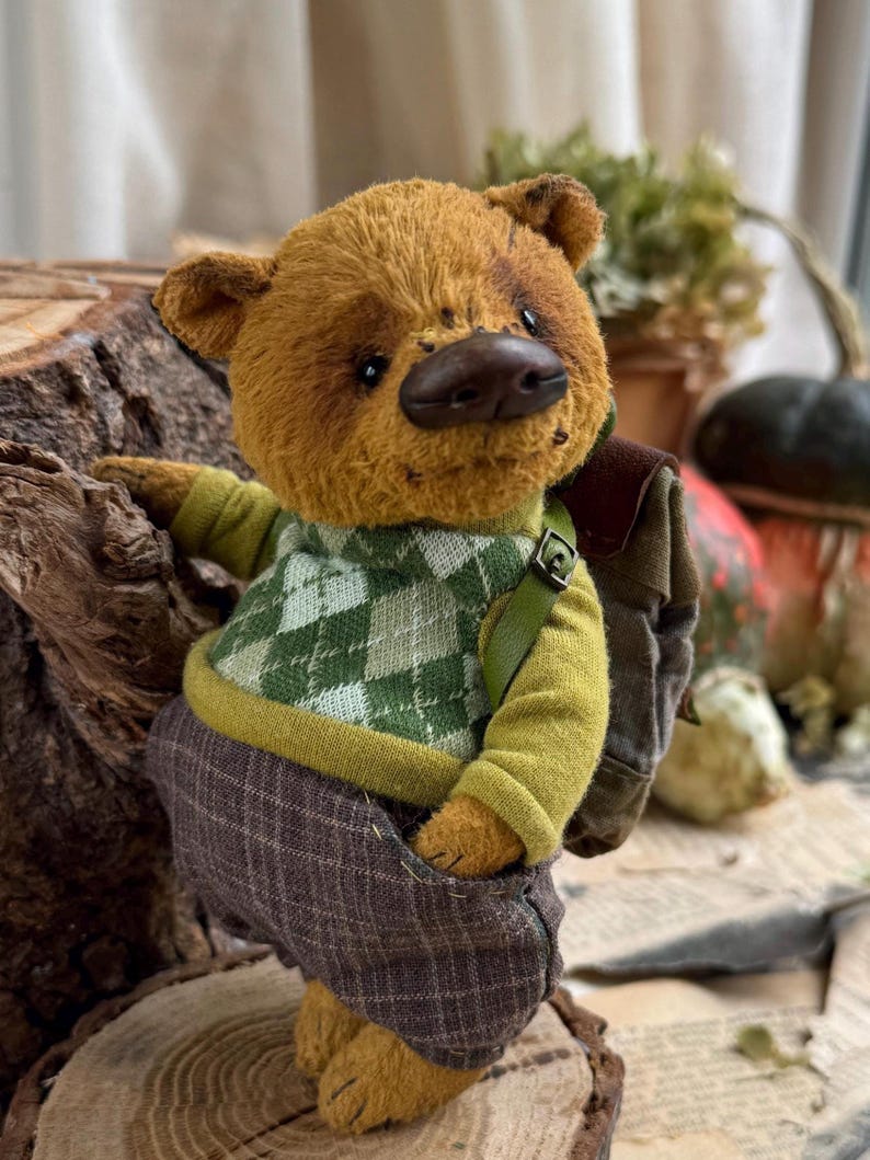 Puede incluir: Un oso de peluche marr&oacute;n que lleva un su&eacute;ter a cuadros verde y blanco, pantalones verdes y una mochila marr&oacute;n. El oso est&aacute; de pie sobre un toc&oacute;n de &aacute;rbol.