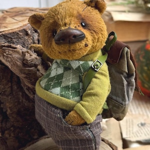 Puede incluir: Un oso de peluche marr&oacute;n que lleva un su&eacute;ter a cuadros verde y blanco, pantalones marrones y una mochila marr&oacute;n. El oso est&aacute; de pie sobre un toc&oacute;n de &aacute;rbol.
