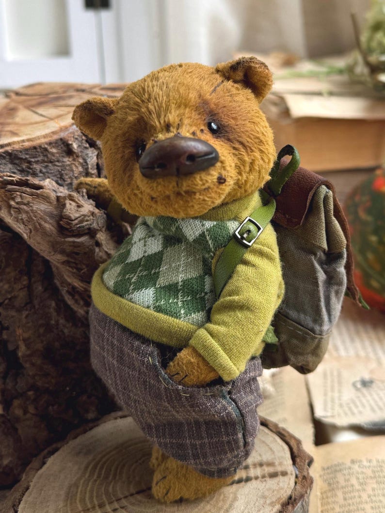 Puede incluir: Un oso de peluche marr&oacute;n que lleva un su&eacute;ter a cuadros verde y blanco, pantalones marrones y una mochila marr&oacute;n. El oso est&aacute; de pie sobre un toc&oacute;n de &aacute;rbol.