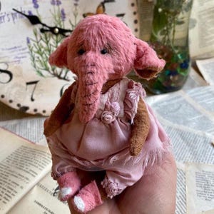 May include: A pink handmade plush elephant wearing a pink dress with lace trim and a floral accent. The elephant has a long trunk and large ears.