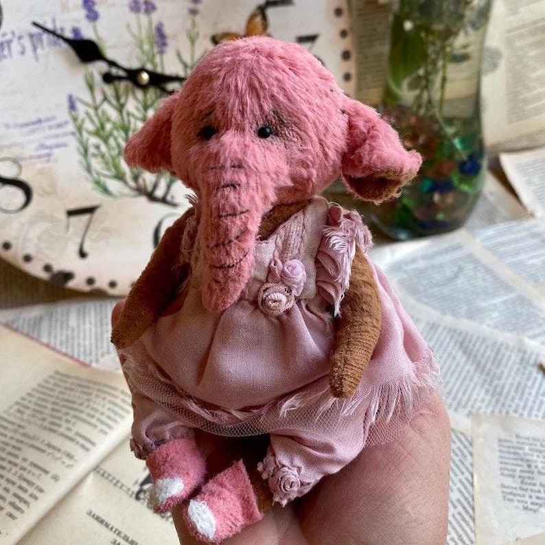 May include: A pink handmade plush elephant wearing a pink dress with lace trim and a floral accent. The elephant has a long trunk and large ears.