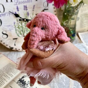 May include: A pink handmade elephant plush toy wearing a pink tutu. The elephant has a brown nose and brown eyes.