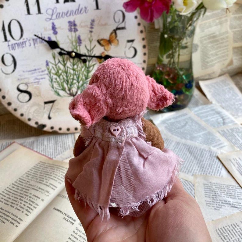 May include: A pink teddy bear wearing a pink dress with a heart-shaped button. The bear is sitting on a hand.