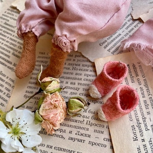 May include: A handmade doll with pink fabric clothing and brown felt boots. The doll is lying on a book with dried roses and a white flower.
