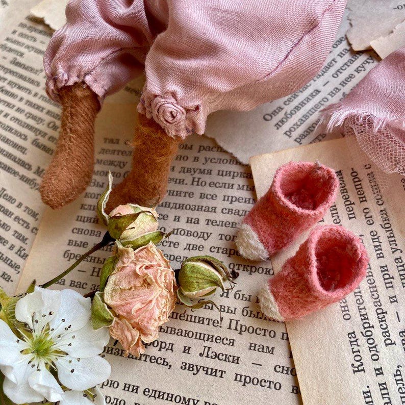 May include: A handmade doll with pink fabric clothing and brown felt boots. The doll is lying on a book with dried roses and a white flower.