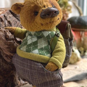 Puede incluir: Un oso de peluche marr&oacute;n que lleva un su&eacute;ter a cuadros verde y blanco, pantalones verdes y una mochila marr&oacute;n. El oso est&aacute; de pie sobre una superficie de madera.