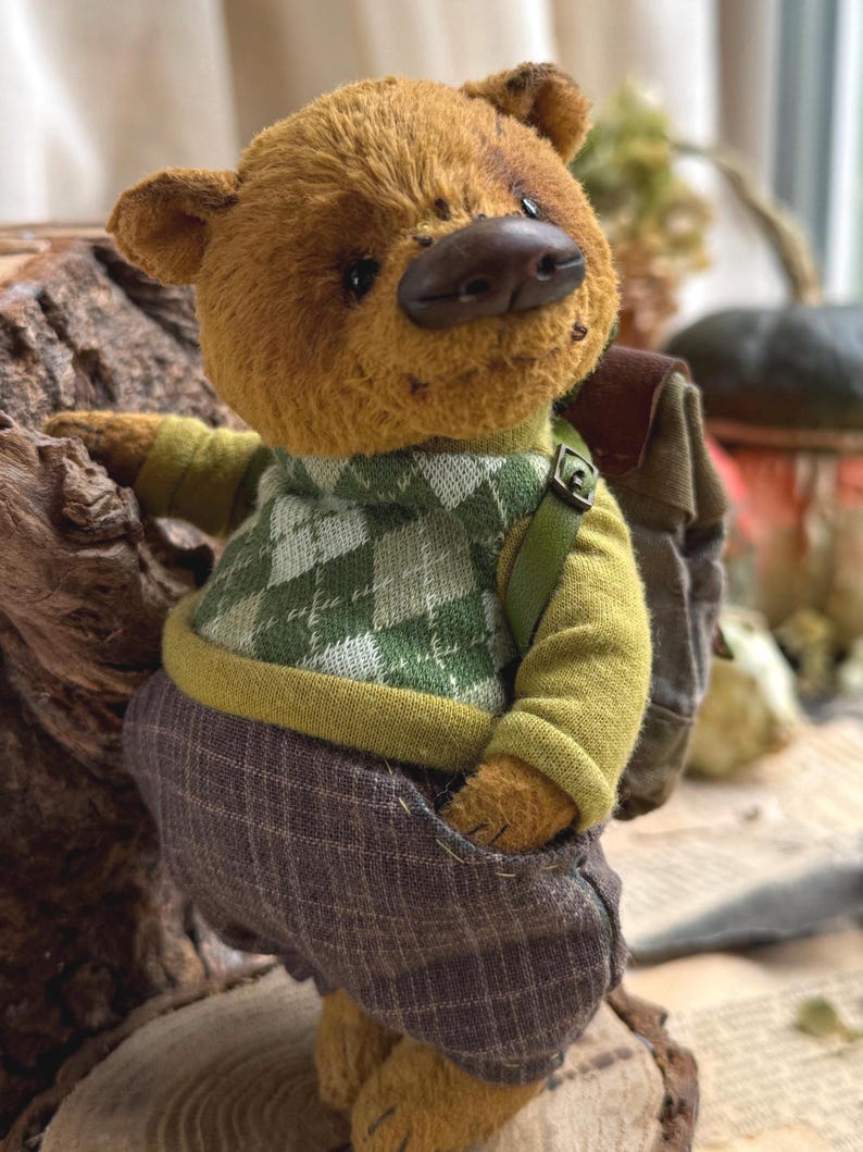 Puede incluir: Un oso de peluche marr&oacute;n que lleva un su&eacute;ter a cuadros verde y blanco, pantalones verdes y una mochila marr&oacute;n. El oso est&aacute; de pie sobre una superficie de madera.