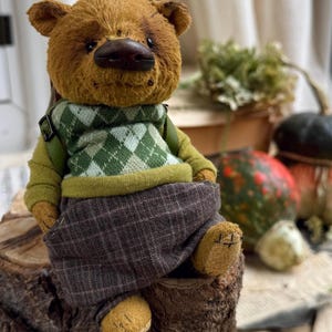 Puede incluir: Un oso de peluche marr&oacute;n con un su&eacute;ter verde y blanco y pantalones de cuadros marrones est&aacute; sentado en un toc&oacute;n de &aacute;rbol. El oso tiene una nariz marr&oacute;n y ojos negros.