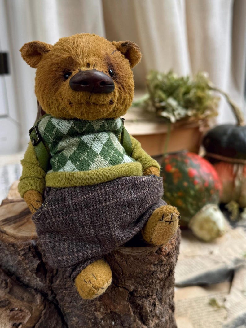 Puede incluir: Un oso de peluche marr&oacute;n con un su&eacute;ter verde y blanco y pantalones de cuadros marrones est&aacute; sentado en un toc&oacute;n de &aacute;rbol. El oso tiene una nariz marr&oacute;n y ojos negros.