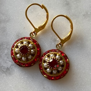 May include: A pair of gold earrings with red and white jeweled accents. The earrings feature a round, red jeweled center surrounded by a ring of white pearls and a ring of smaller red jewels.