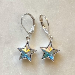 Puede incluir: Un par de pendientes plateados con forma de estrella. Cada pendiente presenta una estrella de cinco puntas con un acabado iridiscente brillante, que muestra tonos de azul, amarillo y verde. Los pendientes tienen un cierre de palanca.
