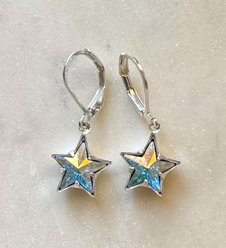 Puede incluir: Un par de pendientes plateados con forma de estrella. Cada pendiente presenta una estrella de cinco puntas con un acabado iridiscente brillante, que muestra tonos de azul, amarillo y verde. Los pendientes tienen un cierre de palanca.