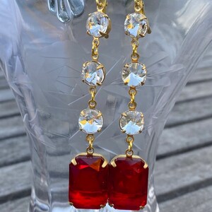 May include: A pair of long gold earrings with red and clear crystal stones. The earrings have a dangling design with multiple clear stones and a large red rectangular stone at the bottom.