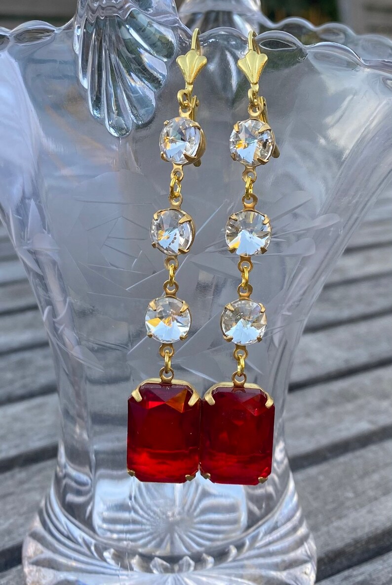 May include: A pair of long gold earrings with red and clear crystal stones. The earrings have a dangling design with multiple clear stones and a large red rectangular stone at the bottom.